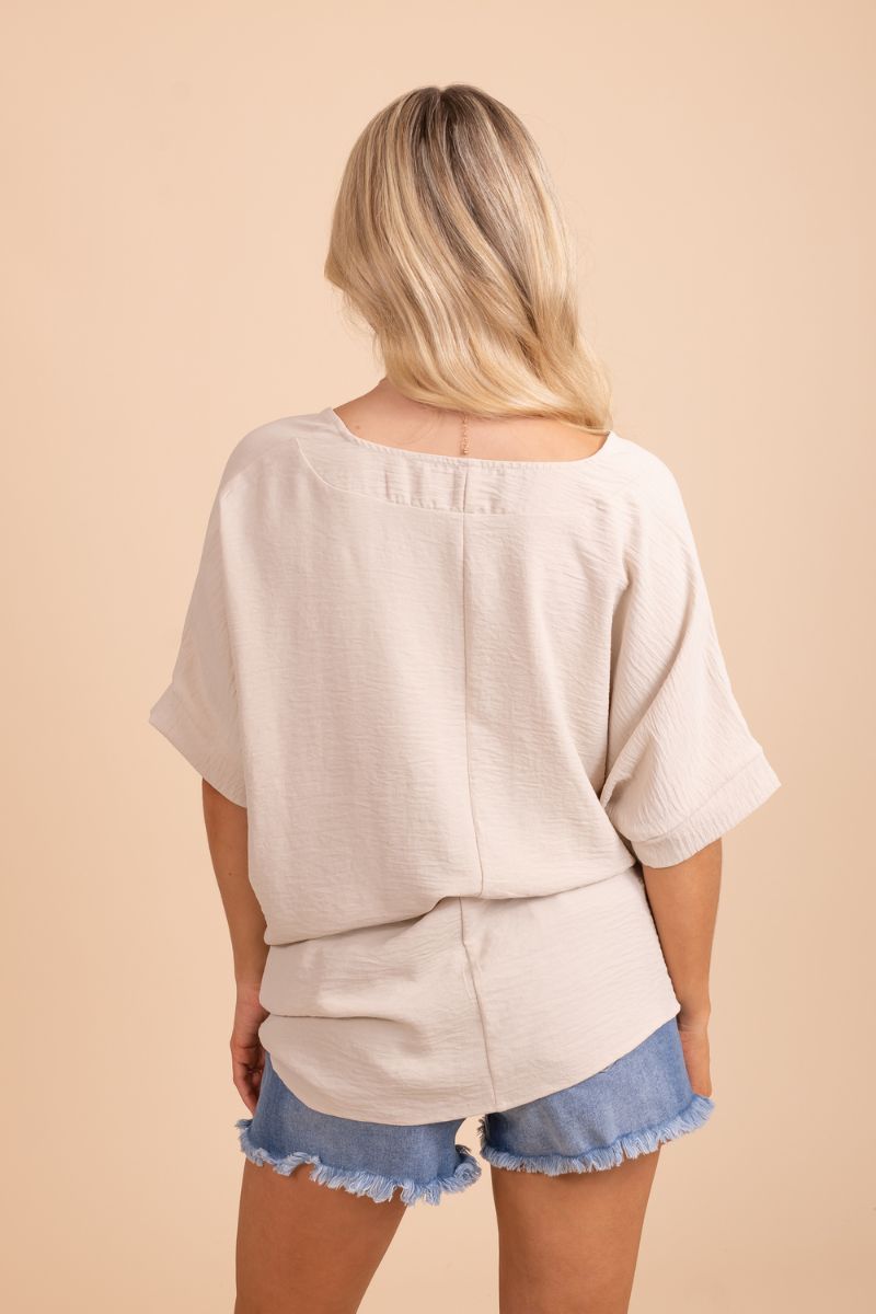 A female model wearing a tan short-sleeve top with a relaxed fit, standing in front of a neutral background.