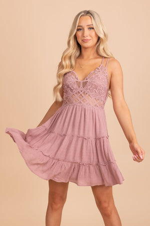 A female model wearing a pink V-neck lace top tiered mini dress, standing in front of a neutral background. She is wearing heels and has her hair styled in loose curls.