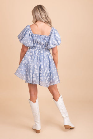 Image of a model wearing a blue puffy dress with white flowers, standing in front of a tan background. The dress has a fitted bodice and a full skirt that falls just above the knee. The blue fabric is covered in white floral patterns. The model is standing with her arms at her side, looking directly at the camera.