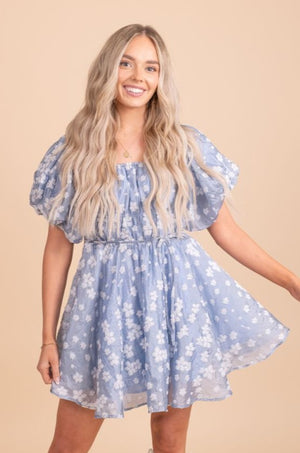 Image of a model wearing a blue puffy dress with white flowers, standing in front of a tan background. The dress has a fitted bodice and a full skirt that falls just above the knee. The blue fabric is covered in white floral patterns. The model is standing with her arms at her side, looking directly at the camera.
