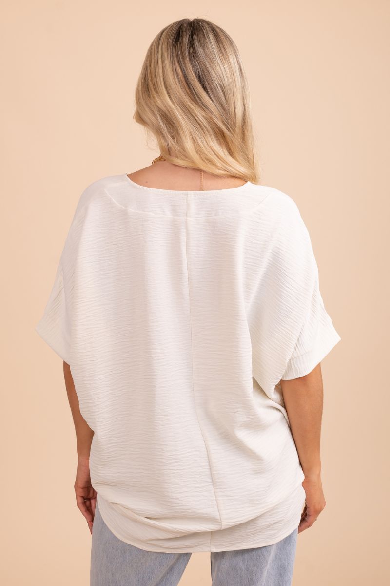 A female model wearing a white flowy top with a subtle V-neck, standing in front of a neutral background.