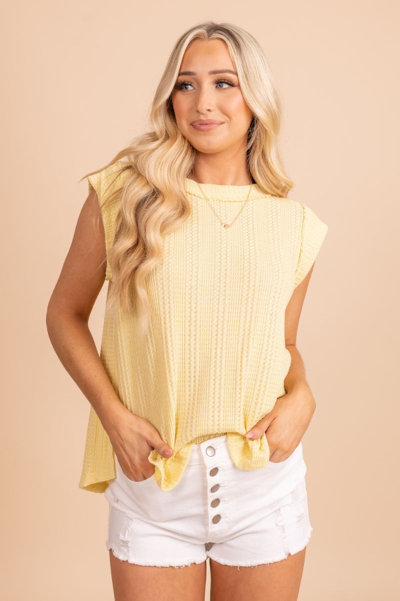 Image of a model wearing a yellow summer top. The top has a flowy and relaxed fit, and the vibrant yellow color is perfect for warm weather. The model is wearing the top with a pair of white shorts and sandals. She is standing in front of a tan background.