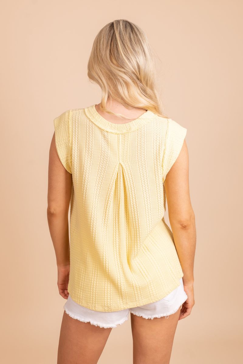 Image of a model wearing a yellow summer top. The top has a flowy and relaxed fit, and the vibrant yellow color is perfect for warm weather. The model is wearing the top with a pair of white shorts and sandals. She is standing in front of a tan background.